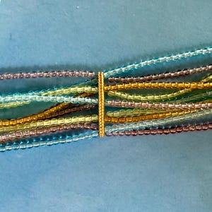 May include: A multi-strand bracelet featuring rows of small, faceted beads in shades of light blue, lavender, lime green, and gold. The strands are held together by two gold-toned metal bars and secured with a matching gold-toned box clasp closure.