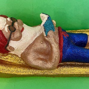 May include: A glittery garden gnome figurine, lying on a gold-colored surface, reading a book. The gnome wears a red hat, sunglasses, blue shorts, and a white shirt. The figurine is a whimsical decorative item.