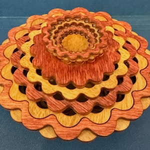 May include: A multi-layered wooden puzzle with a flower-like design. The puzzle features interlocking pieces in shades of red, orange, and yellow. The top layer is a smaller, detailed version of the larger design. The puzzle is made of wood.