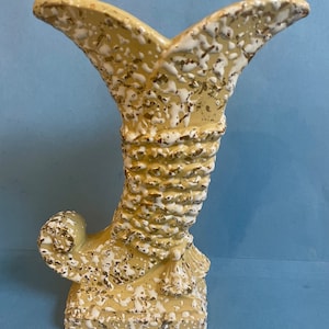May include: A vintage, cream-colored ceramic vase with a textured, speckled finish. The vase has a flared top and a curved, horn-like shape, with white and gold accents. The base is rectangular, and the overall design is reminiscent of a stylized horn.