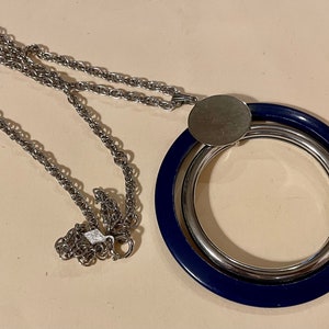 May include: A silver-toned necklace featuring a large pendant with a navy blue outer ring and a silver inner ring. The pendant is connected to a silver chain necklace with a lobster clasp.