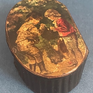 May include: An oval wooden box with a hinged lid. The lid is decorated with a vintage illustration of two children in a forest setting. The children are wearing clothing in shades of brown, red, and white. The box is dark brown and has a ribbed texture.
