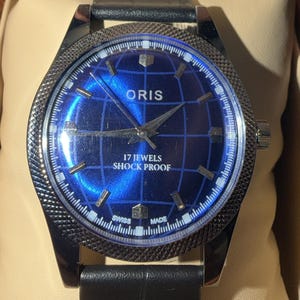 Peut inclure: Une montre-bracelet avec un cadran bleu et un bracelet en cuir noir. Le cadran de la montre présente un motif de carte du monde et le texte "ORIS", "17 JEWELS" et "SHOCK PROOF". La montre est dans une boîte marron.