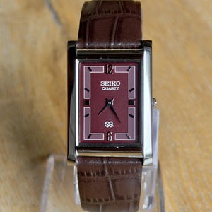May include: A rectangular Seiko Quartz wristwatch featuring a deep burgundy dial with silver-tone hands and markers. The watch has a polished silver-tone metal case and a dark brown leather strap with a textured crocodile-embossed pattern.