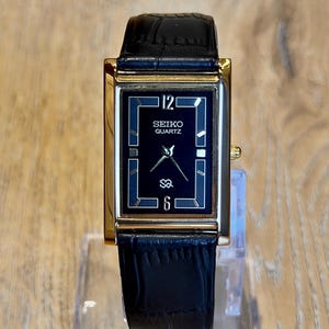 Puede incluir: Un reloj de pulsera rectangular Seiko Quartz con caja dorada, esfera negra con manecillas plateadas y correa de cuero negro con patrón de cocodrilo texturizado. La esfera muestra el número 12 en la parte superior y el 6 en la inferior.