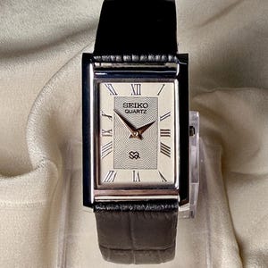 May include: A rectangular Seiko Quartz wristwatch with a silver-toned case and a black leather strap. The watch face features a textured wavy pattern, Roman numeral hour markers, and the Seiko Quartz logo printed in black at the top center.