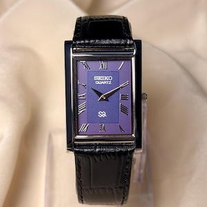 May include: A rectangular Seiko Quartz wristwatch featuring a purple dial with a textured wave pattern, silver-tone hands, and Roman numeral hour markers. The watch has a polished silver-tone case and a black leather strap with a crocodile texture.