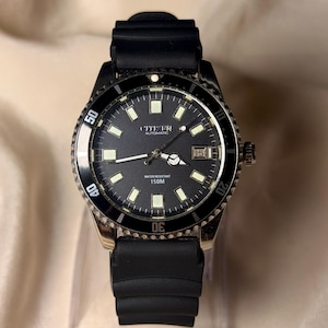 May include: A Citizen automatic wristwatch with a black dial, luminous hour markers, and a date window at the three o'clock position. The watch features a black rotating bezel with white numerals and a black rubber strap with a ribbed texture.