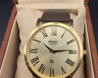 Reloj Seiko de cuarzo vintage: esfera redonda dorada, movimiento japonés, correa de cuero marrón.