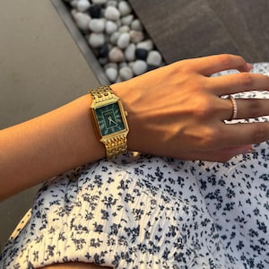 Pu&ograve; includere: Un orologio da polso rettangolare color oro con quadrante verde scuro e numeri romani, dotato di un cinturino a maglie metalliche. L'orologio &egrave; indossato insieme a un sottile anello dorato con piccole pietre chiare su un capo con stampa floreale.