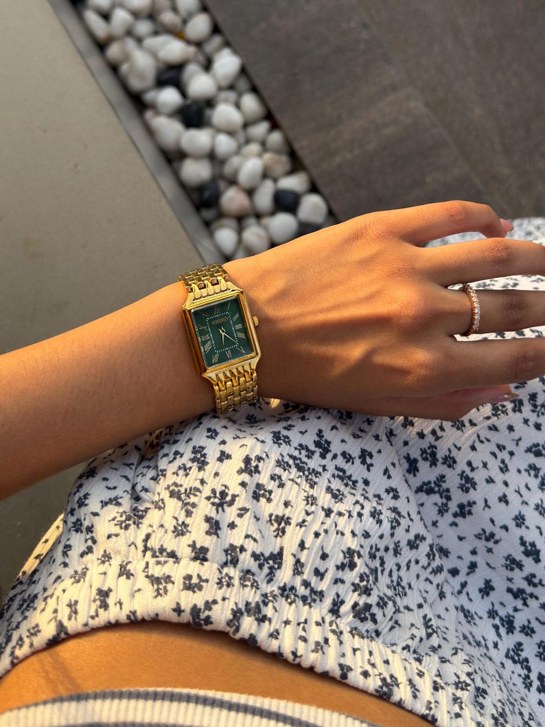 Pu&ograve; includere: Un orologio da polso rettangolare color oro con quadrante verde scuro e numeri romani, dotato di un cinturino a maglie metalliche. L'orologio &egrave; indossato insieme a un sottile anello dorato con piccole pietre chiare su un capo con stampa floreale.