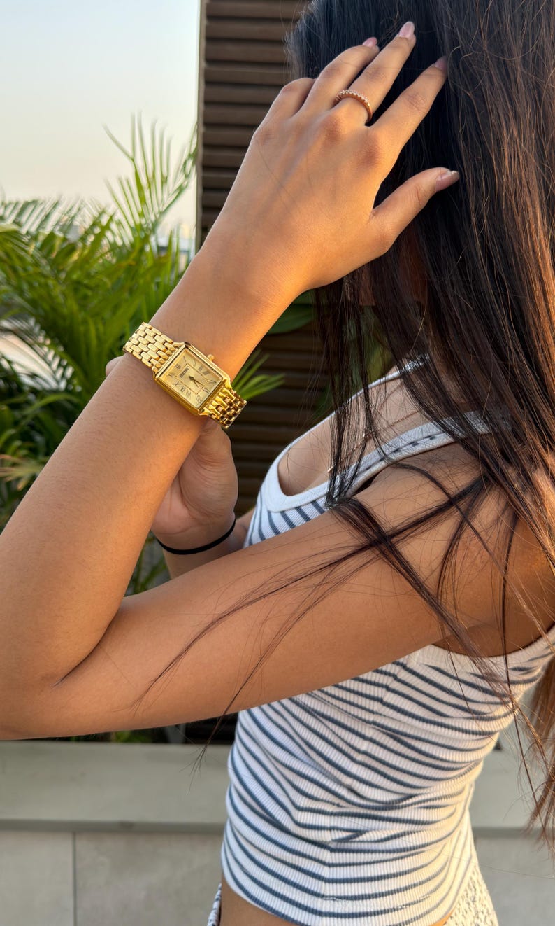Pu&ograve; includere: Un orologio da polso rettangolare color oro con cinturino a maglie metalliche e quadrante chiaro con numeri romani. L'orologio &egrave; indossato al polso e un sottile anello color oro &egrave; visibile su uno delle dita della stessa mano.