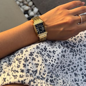 Pu&ograve; includere: Un orologio da polso rettangolare color oro con quadrante nero e numeri romani dorati, dotato di un cinturino a maglie in metallo. Il quadrante mostra il nome del marchio Seiko in lettere dorate sotto la posizione delle ore dodici.