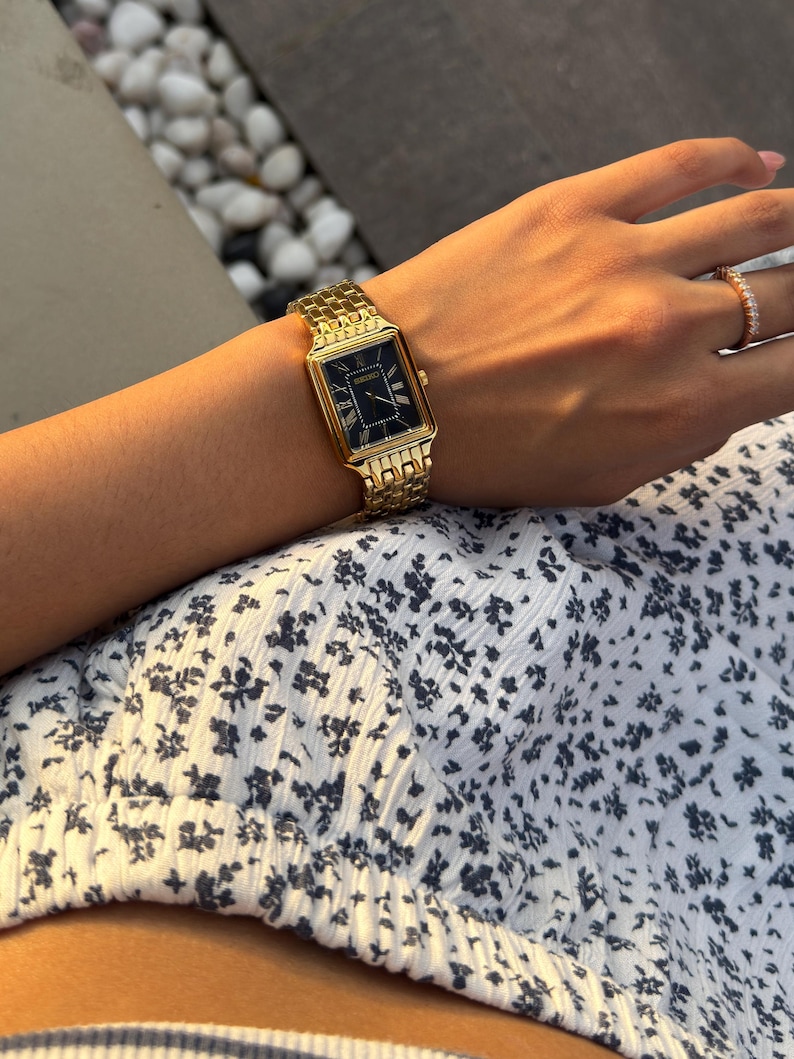 Pu&ograve; includere: Un orologio da polso rettangolare color oro con quadrante nero e numeri romani dorati, dotato di un cinturino a maglie in metallo. Il quadrante mostra il nome del marchio Seiko in lettere dorate sotto la posizione delle ore dodici.