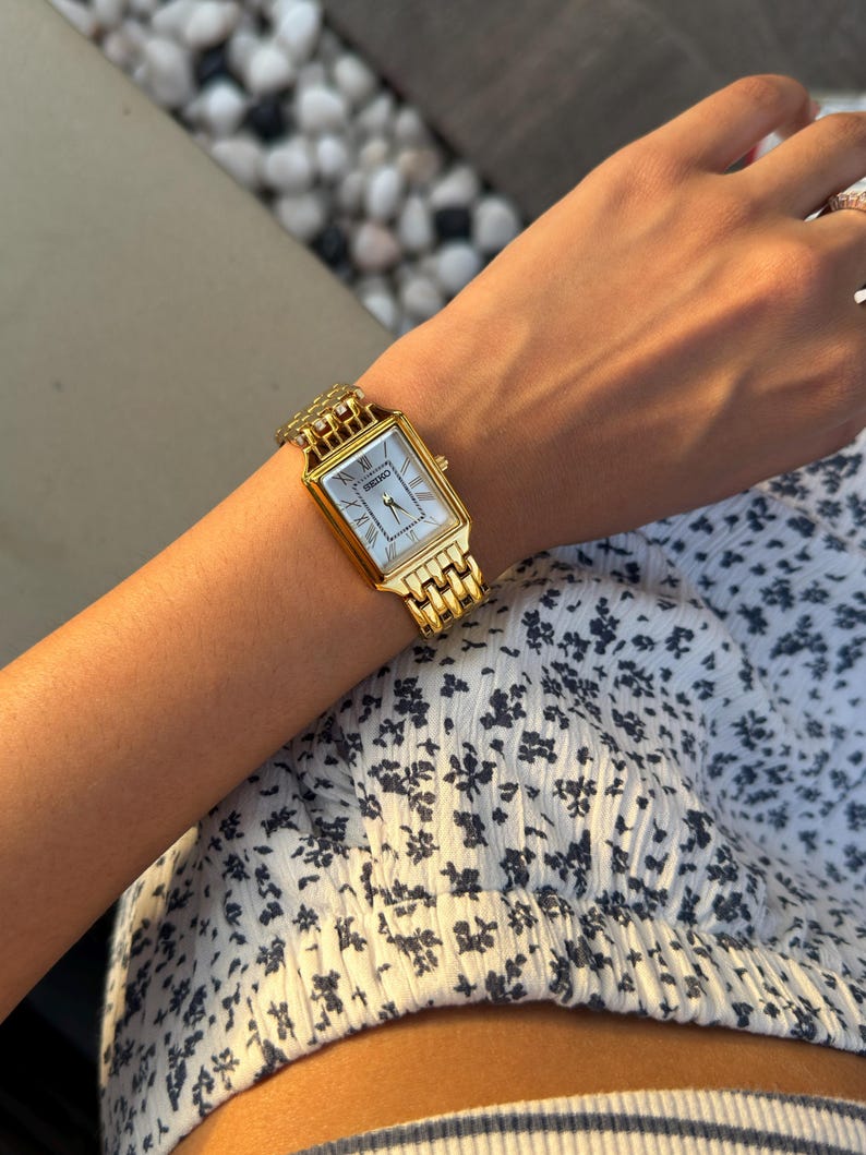 Pu&ograve; includere: Un orologio da polso rettangolare Seiko color oro con quadrante bianco, numeri romani neri come indicatori delle ore e lancette sottili. L'orologio presenta un bracciale a maglie in metallo con una finitura lucida.