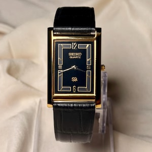 May include: A rectangular Seiko Quartz wristwatch featuring a gold-tone case and a black dial with gold-colored hands and markers. The watch displays the number 12 at the top and 6 at the bottom, with a black leather strap featuring a textured finish.