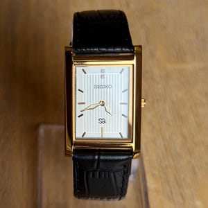 Könnte beinhalten: Eine rechteckige Seiko-Armbanduhr mit goldfarbenem Gehäuse, einem silberfarbenen Zifferblatt mit vertikalen Nadelstreifen, goldfarbenen Zeigern und einem schwarzen Lederarmband mit strukturierter Oberfläche. Das Zifferblatt zeigt das Seiko-Logo.