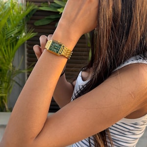 Pu&ograve; includere: Un orologio da polso color oro con cassa rettangolare, quadrante verde scuro e cinturino a maglie in metallo dorato abbinato. L'orologio presenta lancette e indici sottili color oro, offrendo un aspetto chiaro e classico al polso.