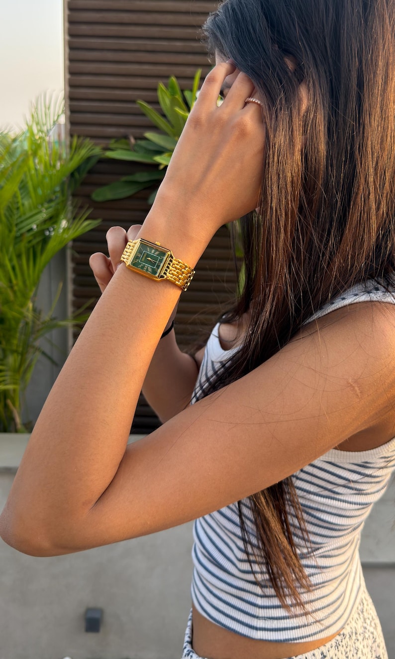 Pu&ograve; includere: Un orologio da polso color oro con cassa rettangolare, quadrante verde scuro e cinturino a maglie in metallo dorato abbinato. L'orologio presenta lancette e indici sottili color oro, offrendo un aspetto chiaro e classico al polso.