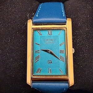 Op de afbeelding: Een rechthoekig, goudkleurig Seiko Quartz horloge met een turquoise wijzerplaat en een blauwe leren band. De wijzerplaat heeft Romeinse cijfers en het Seiko-logo. Het horloge staat tegen een donkere achtergrond.