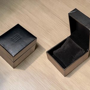 Pu&ograve; includere: Due scatole quadrate nere per gioielli, una chiusa e una aperta. La scatola chiusa presenta un logo sottilmente impresso sul coperchio. La scatola aperta rivela un interno in velluto nero con un morbido cuscino nero per esporre i gioielli.