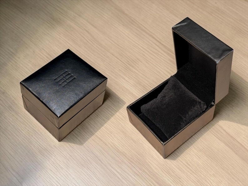 Pu&ograve; includere: Due scatole quadrate nere per gioielli, una chiusa e una aperta. La scatola chiusa presenta un logo sottilmente impresso sul coperchio. La scatola aperta rivela un interno in velluto nero con un morbido cuscino nero per esporre i gioielli.