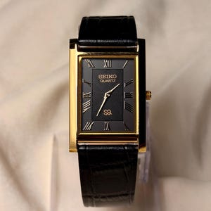 May include: A rectangular Seiko Quartz wristwatch featuring a gold-tone case and a black dial with a textured wave pattern. The dial displays gold Roman numeral hour markers, gold hands, and a black leather strap with a crocodile-embossed texture.