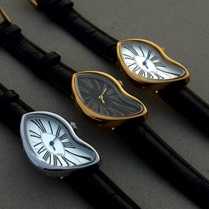 May include: Three wristwatches with irregular, melting-style cases and black leather straps. Two watches feature silver-toned cases with white dials and black Roman numerals, while the center watch has a gold-toned case with a black dial and gold hands.
