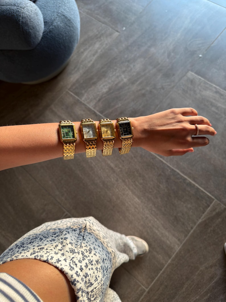 Pu&ograve; includere: Quattro orologi da polso rettangolari con cinturini a maglie in metallo dorato disposti in fila su un braccio. Gli orologi presentano quadranti di diversi colori, tra cui verde, bianco, oro e nero, ciascuno in una cassa dorata coordinata.