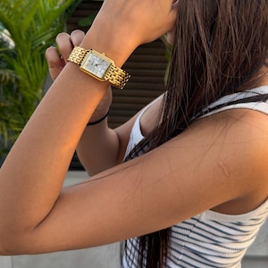 Pu&ograve; includere: Un orologio da polso rettangolare color oro con quadrante bianco e bracciale in metallo a maglie multiple. L'orologio presenta un classico display analogico con lancette e indici neri sottili, fissato da un cinturino in metallo dorato lucido.