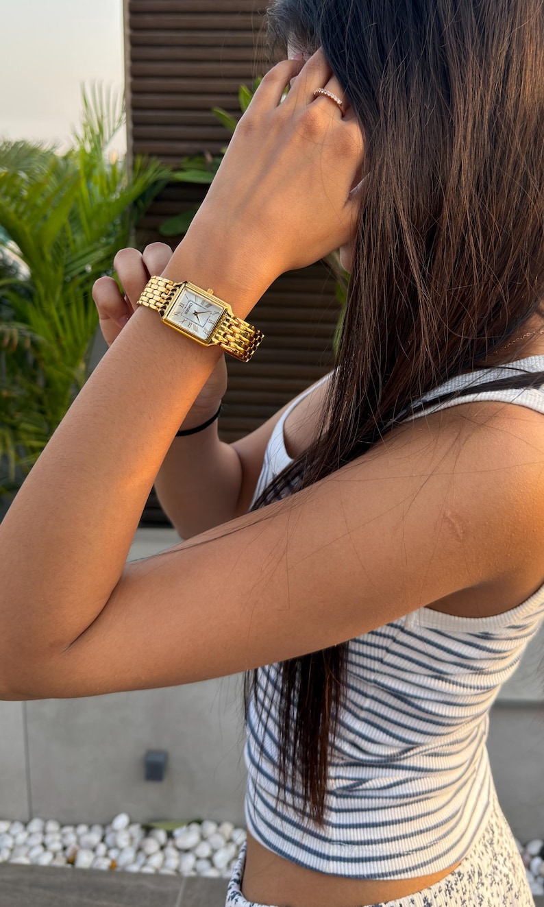 Pu&ograve; includere: Un orologio da polso rettangolare color oro con quadrante bianco e bracciale in metallo a maglie multiple. L'orologio presenta un classico display analogico con lancette e indici neri sottili, fissato da un cinturino in metallo dorato lucido.