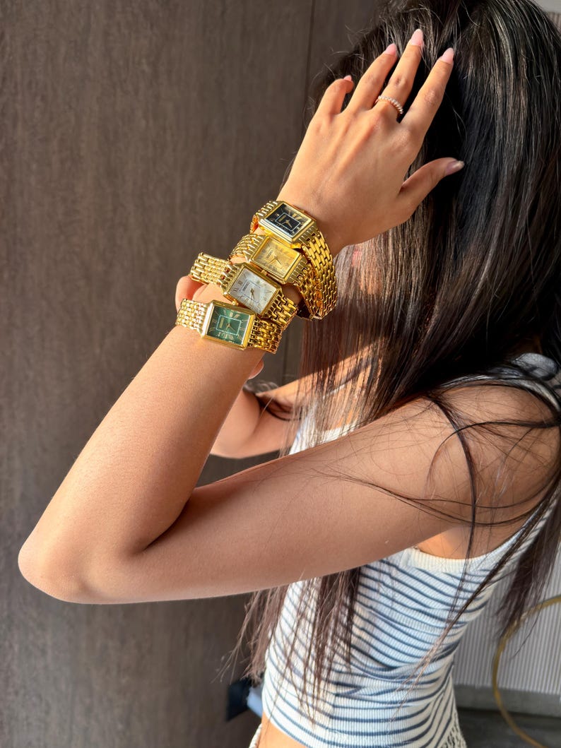 Pu&ograve; includere: Una pila di quattro orologi da polso rettangolari con cinturini a maglie in metallo dorato. Gli orologi presentano quadranti di colori distinti tra cui nero, oro, bianco e verde, ciascuno con indici e lancette dorate, indossati insieme.