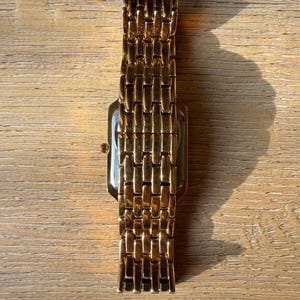 Pu&ograve; includere: Un orologio da polso color oro con cassa rettangolare e cinturino in metallo a maglie multiple. L'orologio presenta una finitura lucida che riflette la luce, con una piccola corona visibile sul lato della cassa.