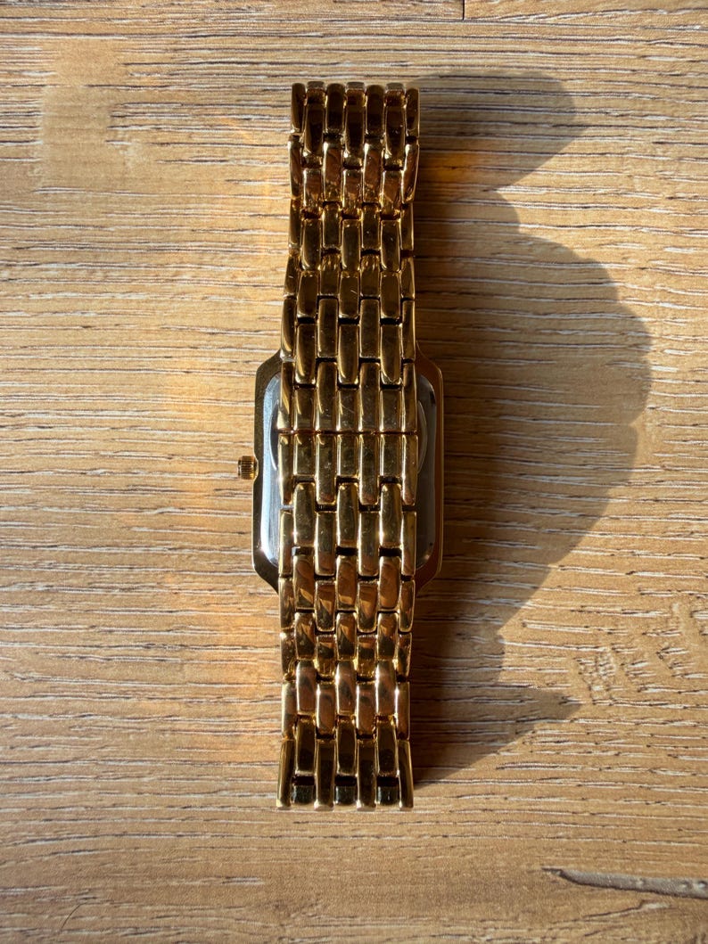 Pu&ograve; includere: Un orologio da polso color oro con cassa rettangolare e cinturino in metallo a maglie multiple. L'orologio presenta una finitura lucida che riflette la luce, con una piccola corona visibile sul lato della cassa.