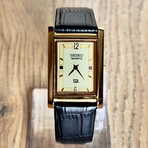 May include: A rectangular Seiko Quartz wristwatch featuring a gold-tone case and a cream-coloured dial with black hands. The watch displays the number 12 at the top and 6 at the bottom, with a black leather strap featuring a textured crocodile pattern.