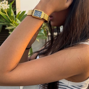 Pu&ograve; includere: Un orologio da polso rettangolare color oro con quadrante nero e numeri romani, dotato di un cinturino a maglie metalliche. L'orologio &egrave; indossato al polso e un sottile anello color oro con piccole pietre &egrave; visibile su un dito della stessa mano.