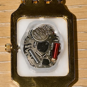 Pu&ograve; includere: Un orologio da polso rettangolare color oro con il fondello rimosso, che rivela il meccanismo interno al quarzo. Il movimento presenta una batteria argentata con etichetta SR626SW, una bobina di rame rossa e la scritta JAPAN UJ20B NO JEWELS.