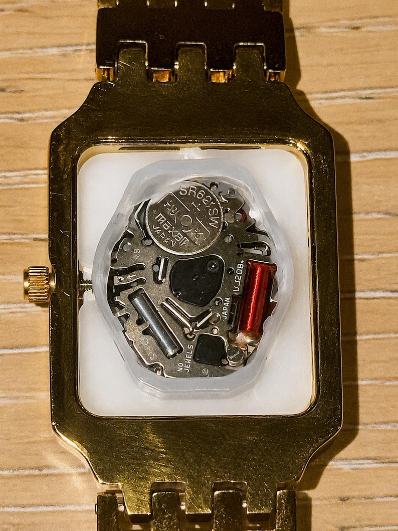 Pu&ograve; includere: Un orologio da polso rettangolare color oro con il fondello rimosso, che rivela il meccanismo interno al quarzo. Il movimento presenta una batteria argentata con etichetta SR626SW, una bobina di rame rossa e la scritta JAPAN UJ20B NO JEWELS.