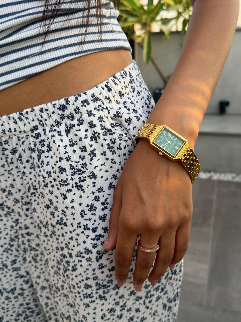 Pu&ograve; includere: Un orologio da polso rettangolare color oro con quadrante verde scuro e numeri romani, dotato di un cinturino a maglie metalliche testurizzate. Un anello sottile e delicato con piccole pietre trasparenti &egrave; indossato all'anulare.