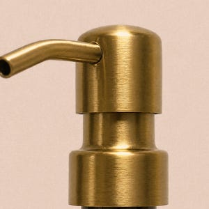 May include: A brushed brass soap dispenser pump with a cylindrical head and a curved, downward-facing spout. The metal finish features a subtle horizontal grain texture, reflecting light across the rounded top and the base of the pump mechanism.