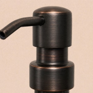 Peut inclure: Une pompe à savon en bronze foncé avec une finition métallique brossée. La tête cylindrique présente un bec verseur incurvé orienté vers le bas pour distribuer le liquide. Le design comprend une section de base circulaire à plusieurs niveaux.