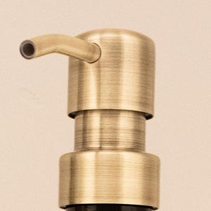 May include: Close-up of a brass soap dispenser pump. The pump has a cylindrical body with a curved spout. The finish has a vintage, antique appearance. The dispenser is designed to be mounted on a bottle.