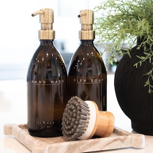 May include: Two amber glass soap dispensers with gold-colored pumps labeled "HAND WASH" and "DISH SOAP" sit on a marble tray. A wooden dish brush with gray bristles rests on the tray. A black vase with greenery is in the background.