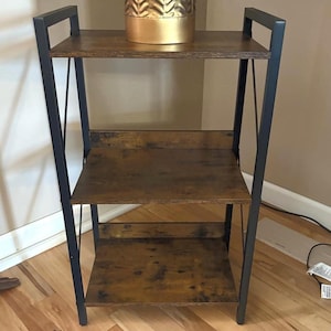 May include: A three-tiered, brown and black bookshelf. The shelves are made of wood with a distressed finish, supported by a black metal frame. The top shelf holds a gold-colored decorative item. The bookshelf is designed for storage and display.