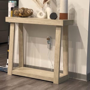 May include: A light-colored wooden console table with a lower shelf. The table is decorated with a gold wire bowl filled with decorative spheres, two white vases, a black disc, and a tall vase with a brown band. Dried ornamental grasses are in one vase.