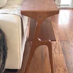 May include: A mid-century modern style wooden side table with a unique, sculptural design. The table has a long, rectangular top and a lower shelf. Two coasters are on the top. The table is made of light brown wood and has a distinctive, curved leg structure.