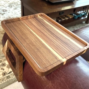 Könnte beinhalten: Ein rechteckiges Sofa-Armtablett aus Holz mit dunkler brauner Oberfläche und einem hellen Streifen. Das Tablett hat einen eingebauten Armlehnenhalter. Das Tablett ist für die Platzierung auf der Armlehne eines Sofas konzipiert.