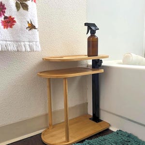 May include: A two-tiered, light-colored bamboo side table with a black metal support. An amber glass spray bottle sits on the top shelf. A floral towel hangs on the wall, and a teal rug is on the floor.