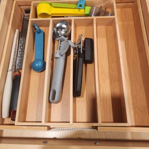 May include: A wooden drawer organizer filled with various kitchen utensils. Items include a grater, knives, a blue scoop, an ice cream scoop, a can opener, and a yellow citrus juicer. The utensils are arranged in separate compartments.