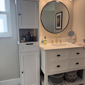 May include: A white bathroom vanity with a sink, drawers, and a lower shelf with two woven baskets. A tall white cabinet stands to the left, and a round mirror hangs above the vanity. A metal bucket with the word "Relax" is on top of the cabinet.